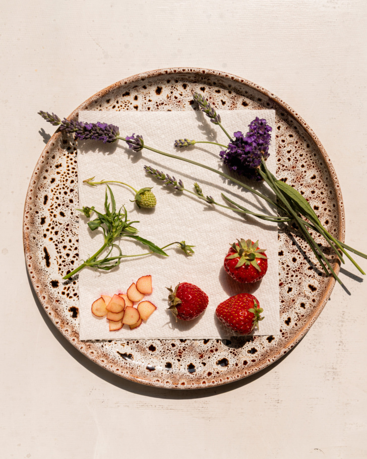An overhead shot shows a speckled plate in direct, warm sunlight that's lined with a paper towel. On top of that is lavender flowers, a tiny green strawberry, 3 small strawberries and razor thin slices of rhubarb.