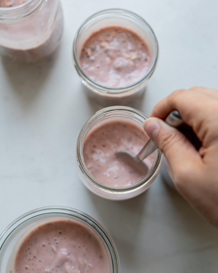An overhead shot shows a hand stirring up some rhubarb strawberry overnight oats in a jar.
