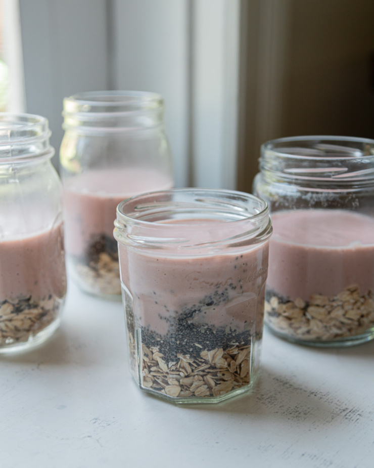 A head-on shot shows jars with layers of oats and seeds and a rhubarb strawberry cashew milk poured on top.