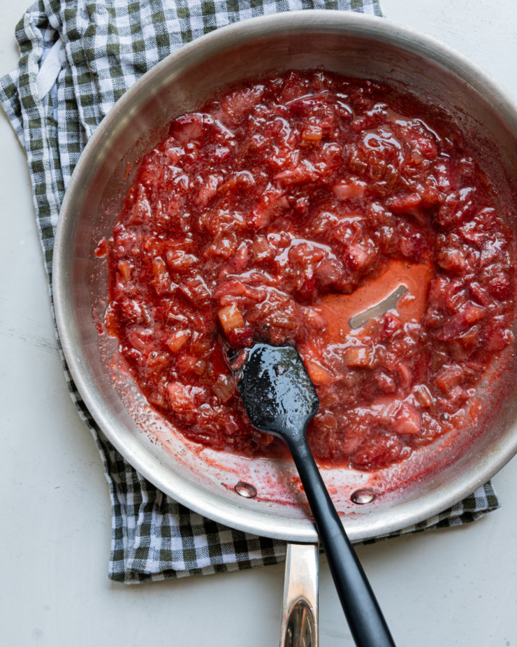 An overhead shot shows a rhubarb strawberry compote in a steel sautรฉ pan with a spatula in the mix.