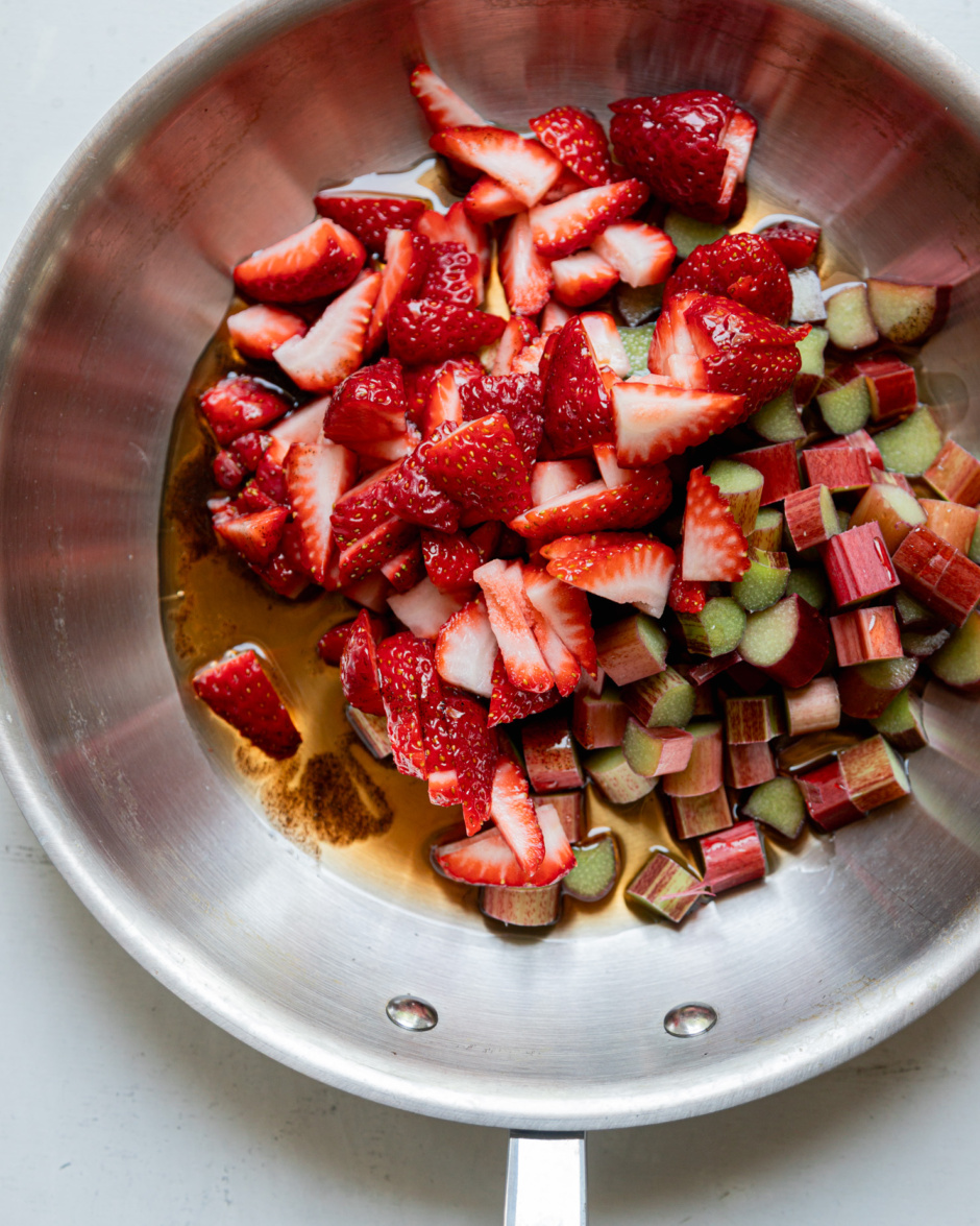 An overhead shot shows chopped rhubarb and strawberries in a sautรฉ pan with maple syrup and vanilla bean paste.