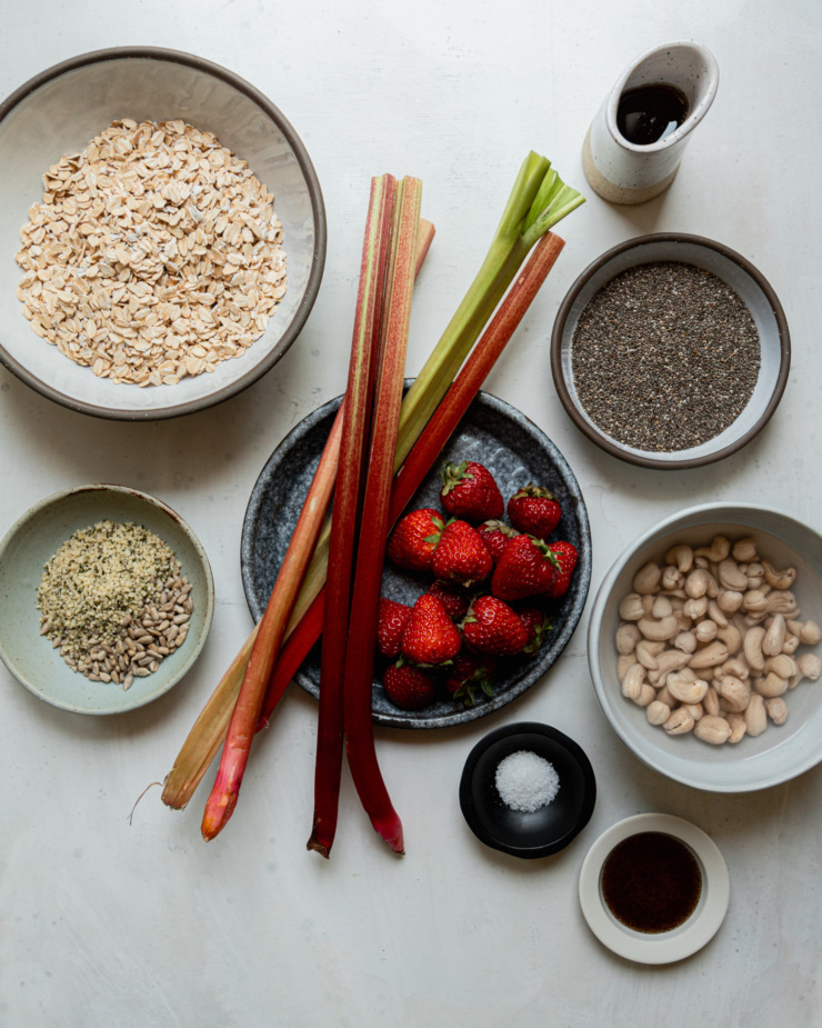 An overhead shot shows measured out ingredients for rhubarb strawberry overnight oats: rolled oats, rhubarb stalks, strawberries, maple syrup, chia seeds, raw cashews, vanilla bean paste, salt, hemp, and sunflower seeds.