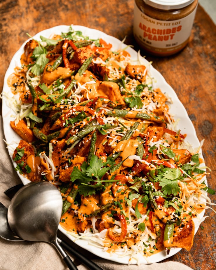 A 3/4 angle shot shows a Gochujang peanut sweet potato salad with green beans, red bell peppers, shredded cabbage, and chopped herbs. The roasted vegetables are piled on top of the cabbage on an oval platter and topped with peanut Gochujang dressing and sesame seeds. A jar of Logan Petit Lot peanut butter and serving tongs are seen nearby.