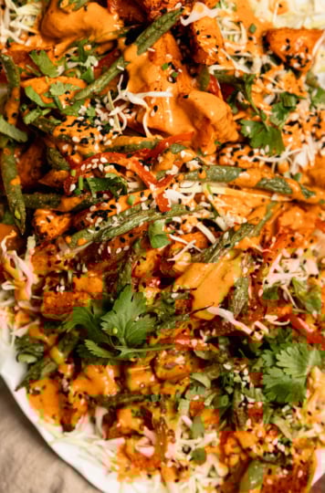 An up close, overhead shot shows a Gochujang peanut sweet potato salad with green beans, red bell peppers, shredded cabbage, and chopped herbs. The roasted vegetables are piled on top of the cabbage on an oval platter and topped with peanut Gochujang dressing and sesame seeds.