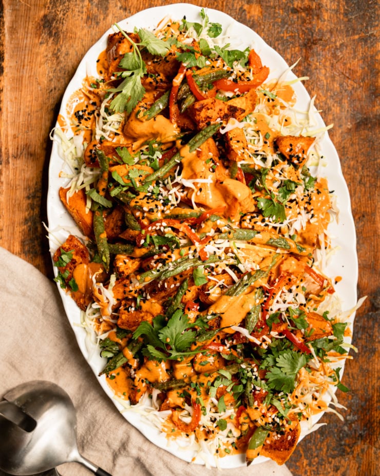 An overhead shot shows a Gochujang peanut sweet potato salad with green beans, red bell peppers, shredded cabbage, and chopped herbs. The roasted vegetables are piled on top of the cabbage on an oval platter and topped with peanut Gochujang dressing and sesame seeds.
