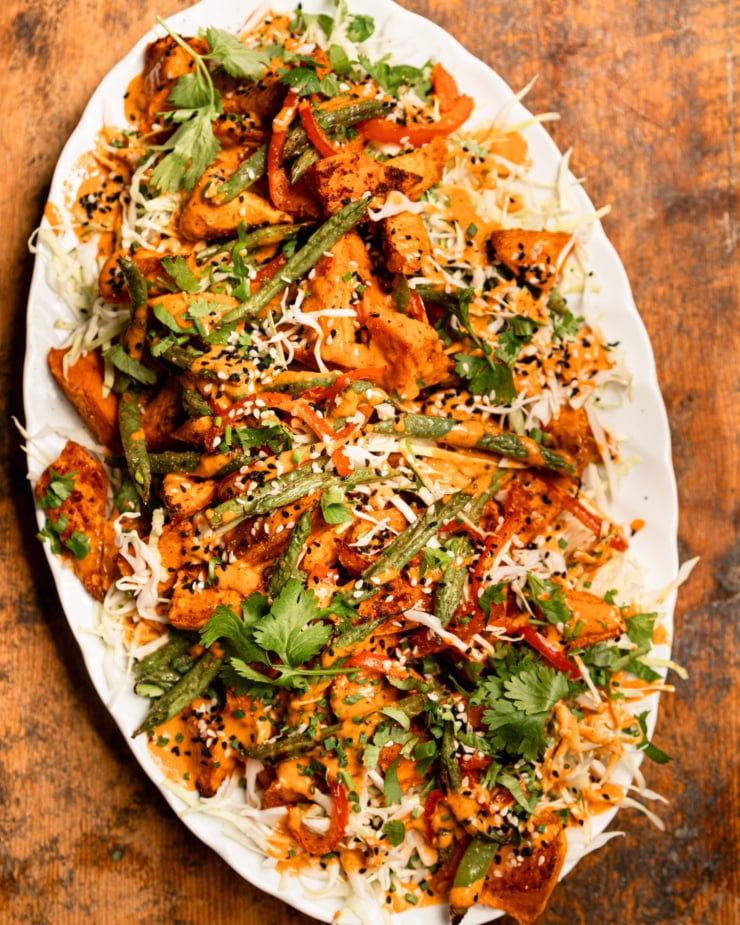 An overhead shot shows a Gochujang peanut sweet potato salad with green beans, red bell peppers, shredded cabbage, and chopped herbs. The roasted vegetables are piled on top of the cabbage on an oval platter and topped with peanut Gochujang dressing and sesame seeds.