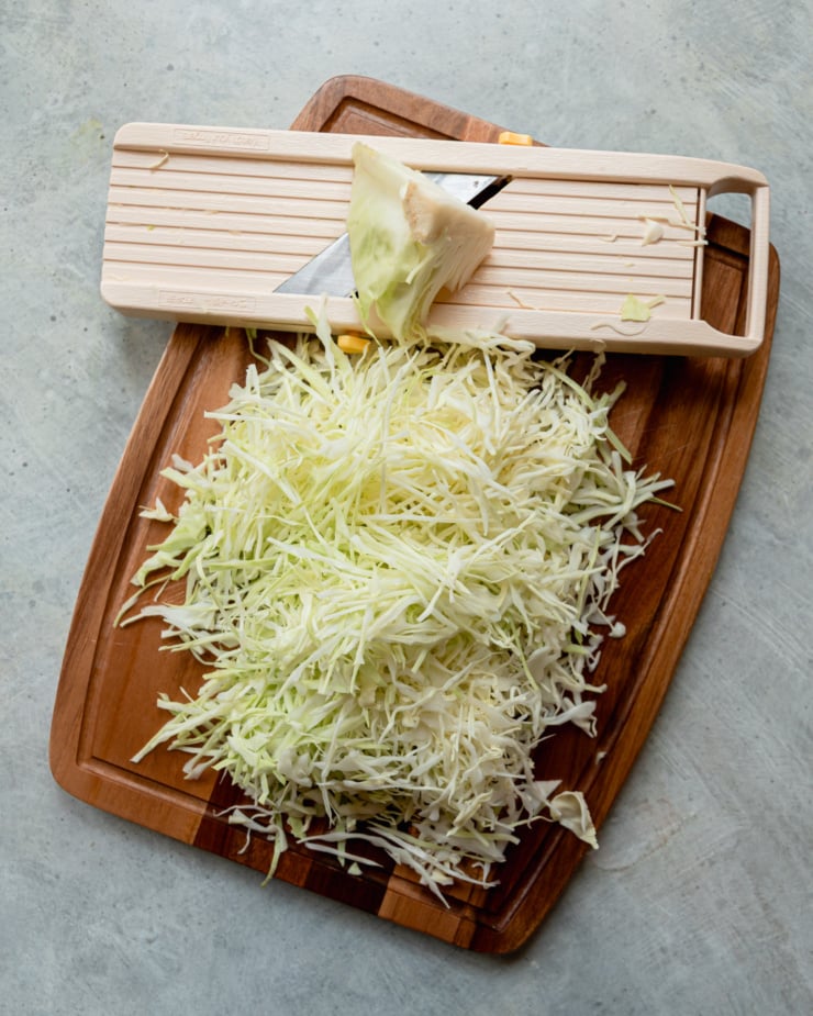 An overhead shot shows a cutting board with a bunch of shredded cabbage and a mandoline slicer on top.