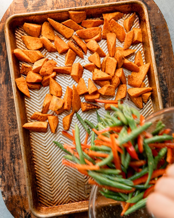 An overhead shot shows roasted sweet potatoes on a baking sheet. Green beans and red bell pepper strips are being emptied onto the baking sheet from a bowl.