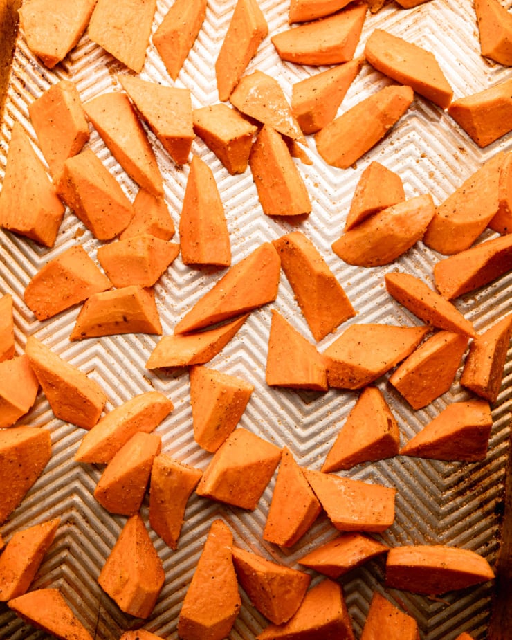 An overhead shot shows 2-inch pieces of sweet potato on a baking sheet, coated in oil and spices.