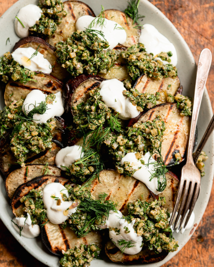 An up close, overhead shot shows a platter of grilled Japanese sweet potato slices topped with dollops of dill pickle salsa verde, dollops of vegan sour cream, and fresh dill.