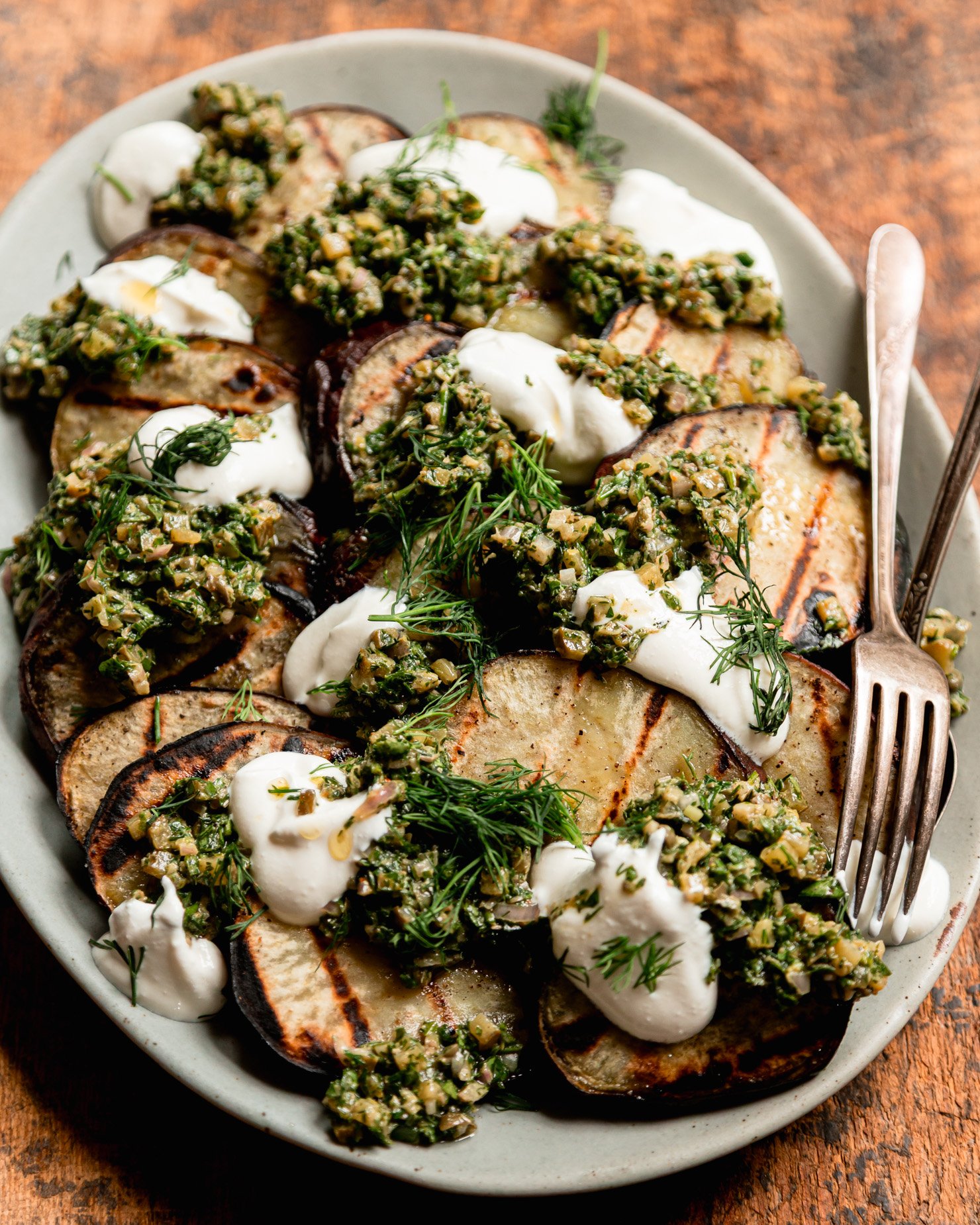 A 3/4 angle shot shows a platter of grilled Japanese sweet potato slices topped with dollops of dill pickle salsa verde, dollops of vegan sour cream, and fresh dill.