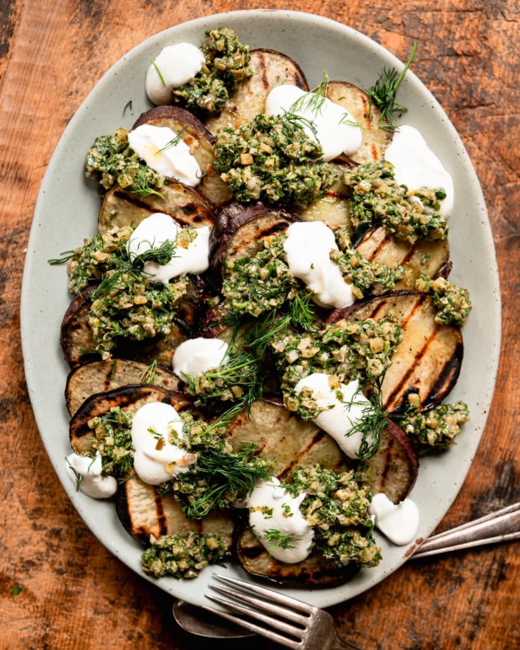 An overhead shot shows a platter of grilled Japanese sweet potato slices topped with dollops of dill pickle salsa verde, dollops of vegan sour cream, and fresh dill.