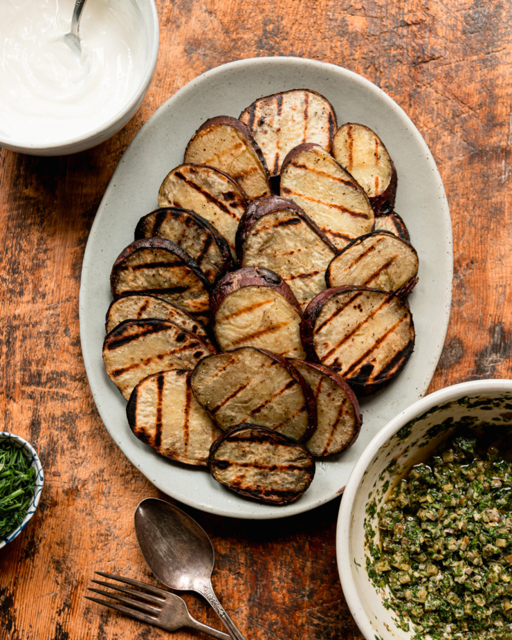An overhead shot shows a platter of grilled Japanese sweet potato slices, a bowl of dill pickle salsa verde, and a bowl of vegan sour cream.