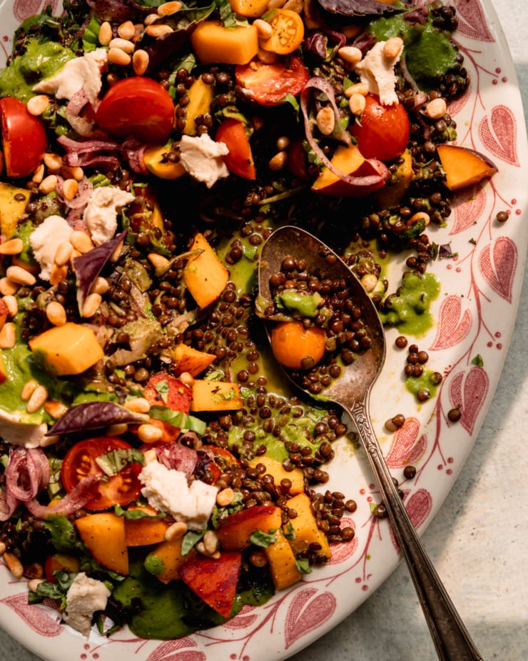 An up close, overhead shot shows a black lentil salad on a platter with diced peaches, cherry tomatoes, vegan feta, sumac onions, and basil vinaigrette. A serving spoon is perched on the side of the platter after having scooped out some of the salad.