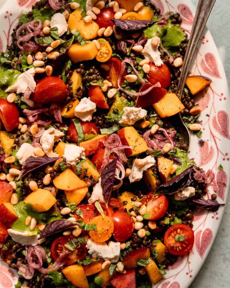 An up close, overhead shot shows a black lentil salad on a platter with diced peaches, cherry tomatoes, vegan feta, sumac onions, pine nuts, and basil vinaigrette. A serving spoon is tucked into the side of the salad.