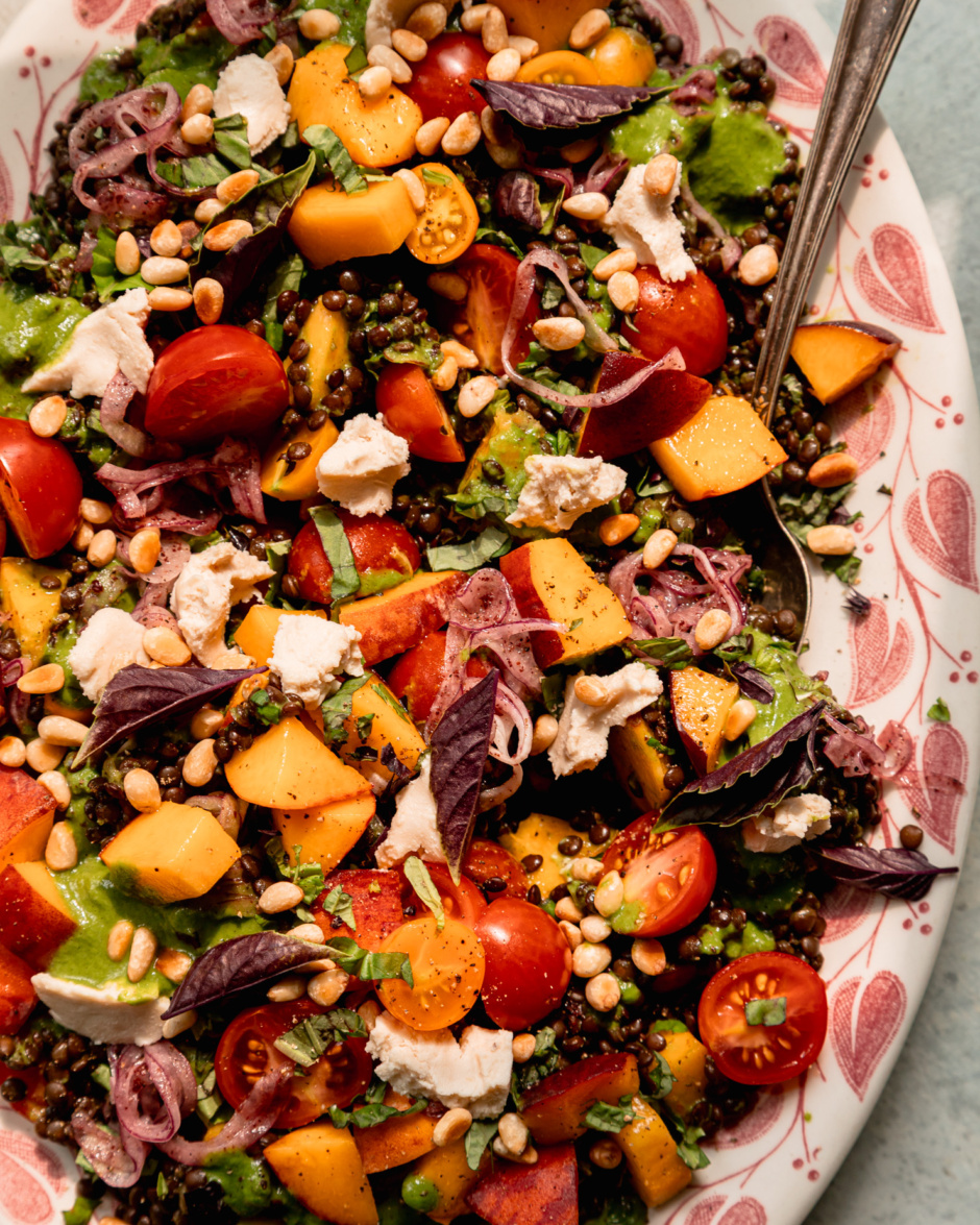 An up close, overhead shot shows a black lentil salad on a platter with diced peaches, cherry tomatoes, vegan feta, sumac onions, pine nuts, and basil vinaigrette. A serving spoon is tucked into the side of the salad.