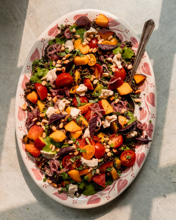 An overhead shot shows a black lentil salad on a platter with diced peaches, cherry tomatoes, vegan feta, sumac onions, pine nuts, and basil vinaigrette. A serving spoon is tucked into the side of the salad. The photo is shot in direct sun light.