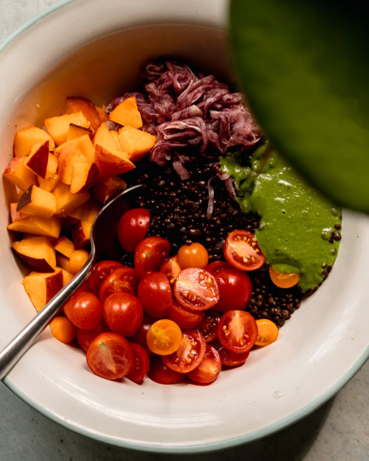 An overhead shot shows a bowl with cooked black lentils, diced peaches, halved cherry tomatoes, and thinly sliced sumac onions. A green and creamy basil vinaigrette is being poured over top.