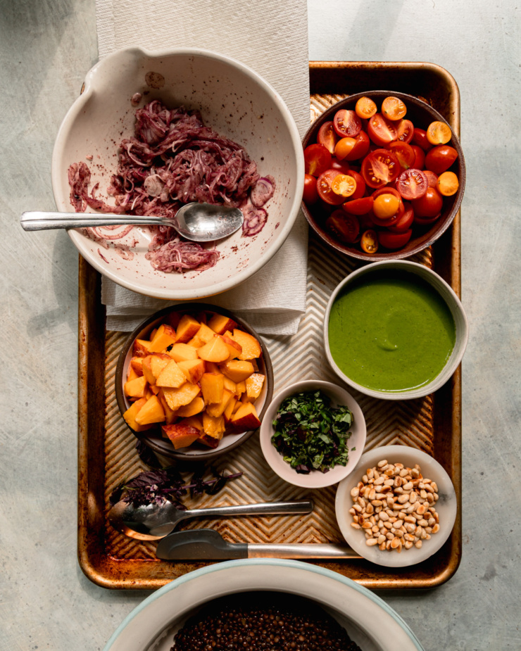 An overhead shot shows a bowl of cooked black lentils as well as a baking sheet with the following in prep bowls: sumac onions, halved cherry tomatoes, diced peaches, chopped basil, toasted pine nuts, and basil vinaigrette.