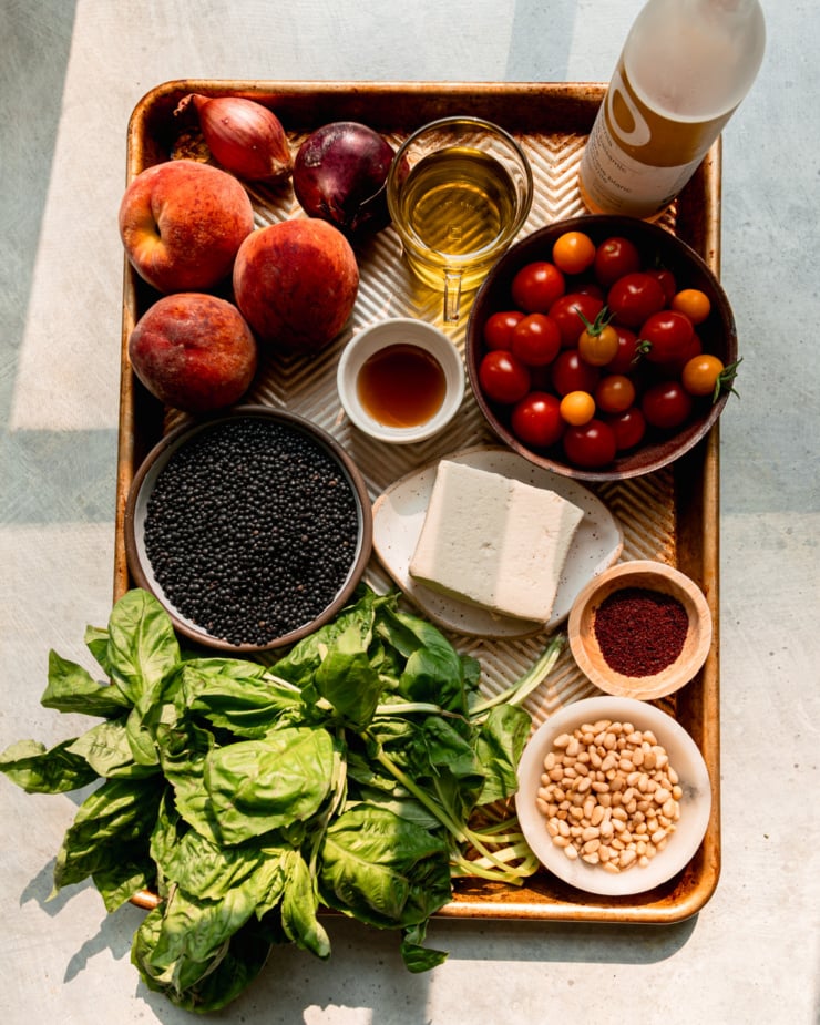 An overhead shot shows ingredients for a black lentil salad: olive oil, white balsamic vinegar, cherry tomatoes, agave nectar, vegan feta, sumac, pine nuts, basil, black lentils, peaches, a shallot, and a red onion. Everything is contained on a baking sheet in bright sun light.