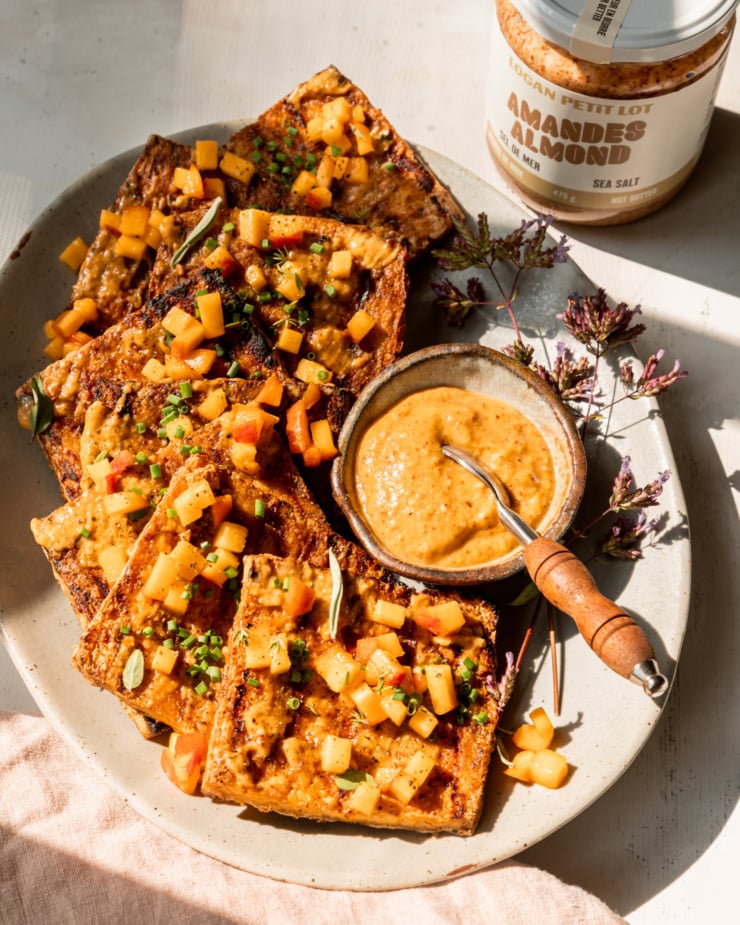A 3/4 angle shot shows a platter with grilled tofu slices, brushed with peach, almond butter & miso glaze. The tofu is garnished with tiny dices of peach and chopped herbs. A small bowl of the sauce with a small spoon is seen on the side of the plate. The photo is taken in direct sun light with some harsh shadows. A jar of Logan Petit Lot almond butter is seen to the side.