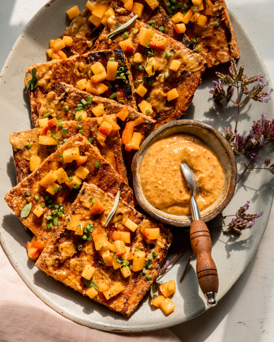 An overhead shot shows a platter with grilled tofu slices, brushed with peach, almond butter & miso glaze. The tofu is garnished with tiny dices of peach and chopped herbs. A small bowl of the sauce with a small spoon is seen on the side of the plate. The photo is taken in direct sun light with some harsh shadows.