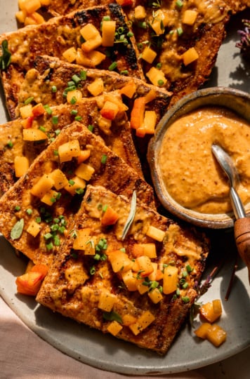 An up close, overhead shot shows a platter with grilled tofu slices, brushed with peach, almond butter & miso glaze. The tofu is garnished with tiny dices of peach and chopped herbs. A small bowl of the sauce with a small spoon is seen on the side of the plate. The photo is taken in direct sun light with some harsh shadows.
