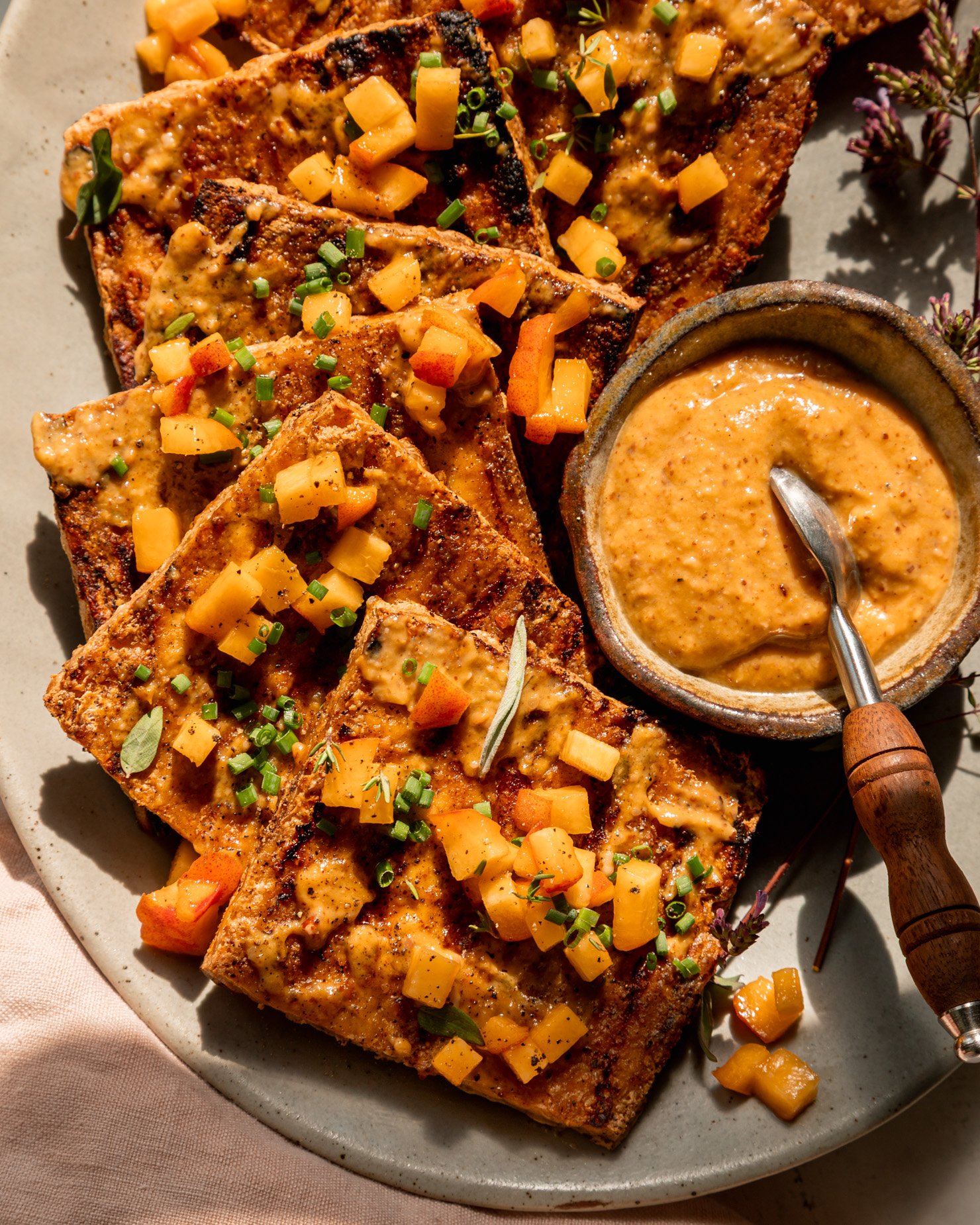 An up close, overhead shot shows a platter with grilled tofu slices, brushed with peach, almond butter & miso glaze. The tofu is garnished with tiny dices of peach and chopped herbs. A small bowl of the sauce with a small spoon is seen on the side of the plate. The photo is taken in direct sun light with some harsh shadows.