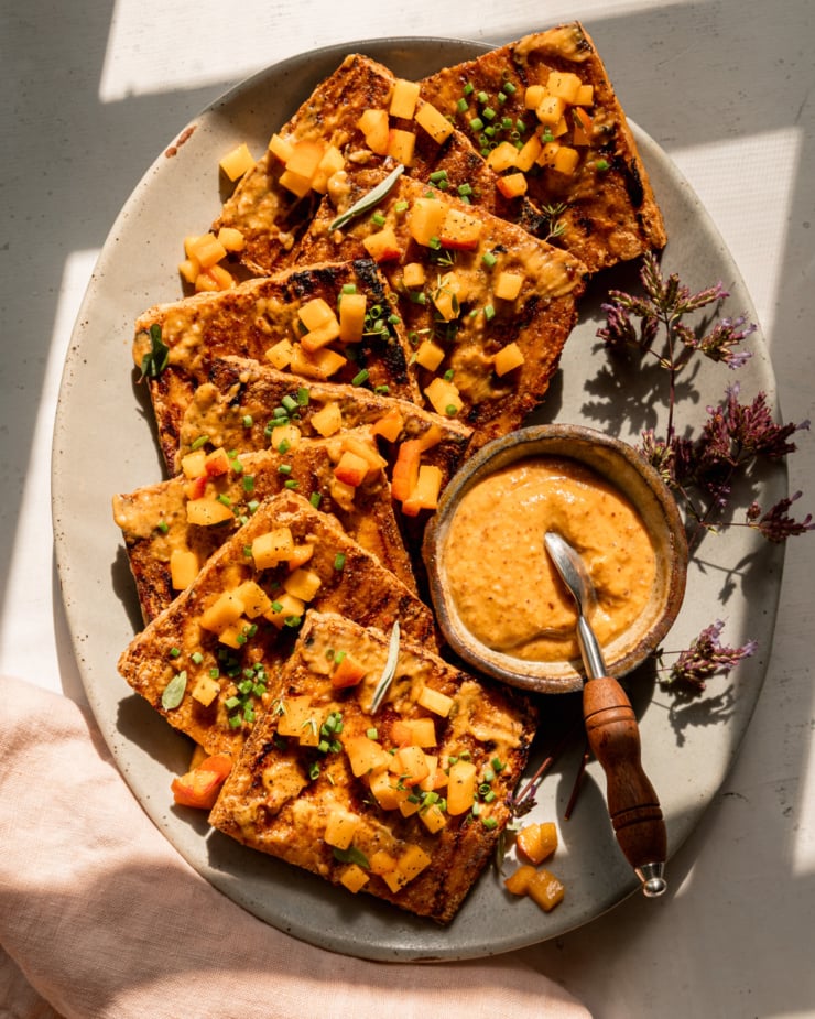 An overhead shot shows a platter with grilled tofu slices, brushed with peach, almond butter & miso glaze. The tofu is garnished with tiny dices of peach and chopped herbs. A small bowl of the sauce with a small spoon is seen on the side of the plate. The photo is taken in direct sun light with some harsh shadows.
