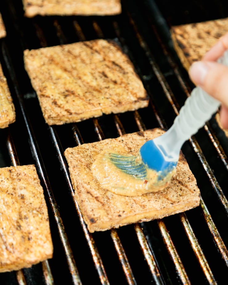 A 3/4 angle image shows grilled tofu slices on a grill. One slice is being brushed with a creamy sauce.