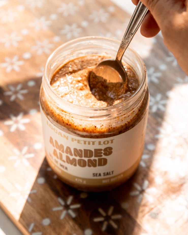 An overhead shot shows a hand using a spoon to stir up a jar of almond butter in bright sun light.
