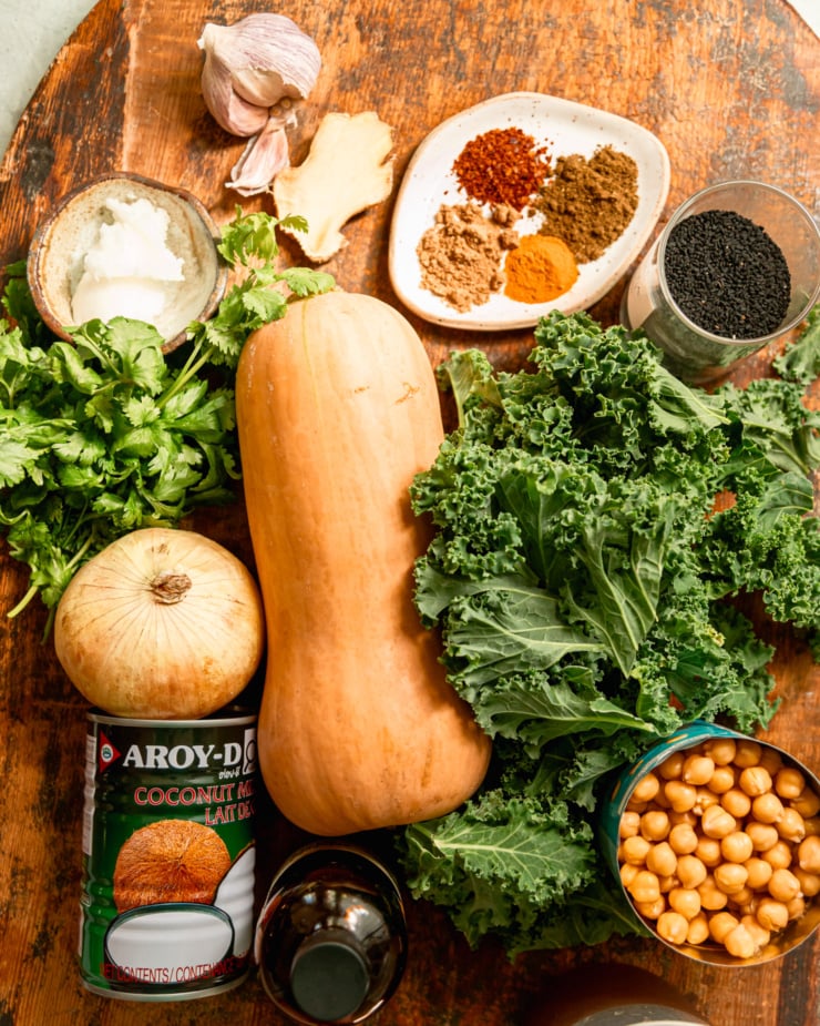 An overhead shot shows ingredients needed for a butternut squash stew with chickpeas and kale. All ingredients are on a rough wood board.