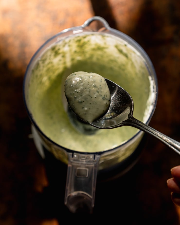 An overhead shot shows a spoon of creamy green-flecked cilantro sauce hovering over a mini food processor filled with the same sauce.