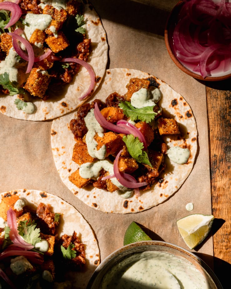 A overhead image shows 3 vegan butternut squash tacos with creamy cilantro sauce drizzled on top. The tacos also feature a smashed chipotle pinto bean base, chopped cilantro, and pickled red onions. Lime wedges and a bowl of the sauce are seen nearby.