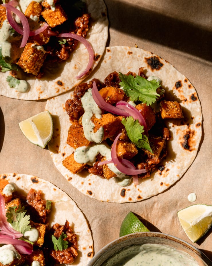 An up close, overhead image shows 3 vegan butternut squash tacos with creamy cilantro sauce drizzled on top. The tacos also feature a smashed chipotle pinto bean base, chopped cilantro, and pickled red onions. Lime wedges are seen nearby.