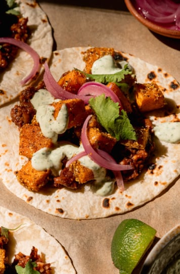 A 3/4 angle image shows a vegan butternut squash taco with creamy cilantro sauce drizzled on top. The taco also features a smashed chipotle pinto bean base, chopped cilantro, and pickled red onions. A lime wedge is seen nearby.