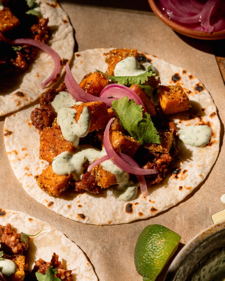 A 3/4 angle image shows a vegan butternut squash taco with creamy cilantro sauce drizzled on top. The taco also features a smashed chipotle pinto bean base, chopped cilantro, and pickled red onions. A lime wedge is seen nearby.