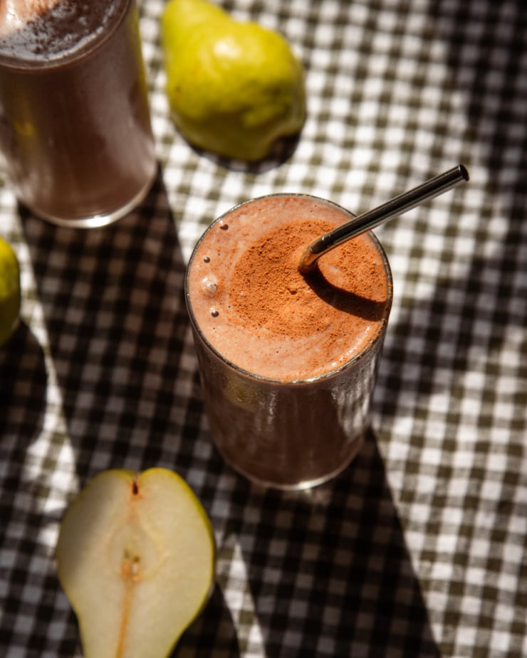 An overhead shot shows a glass of chocolate pear smoothie on top of a checked napkin. The smoothie is garnished with a dusting of cinnamon and a straw is sticking out.