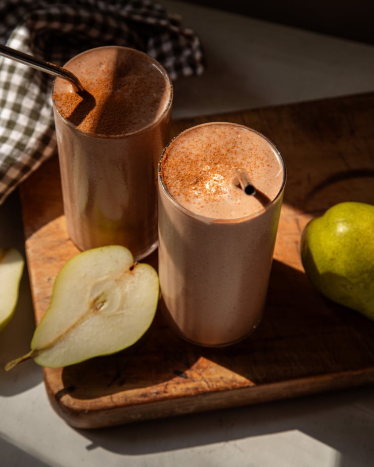 An overhead shot shows two chocolate pear smoothies on a wood board in bright sun light. They have cinnamon sprinkled on top and stainless straws sticking out of the glasses.