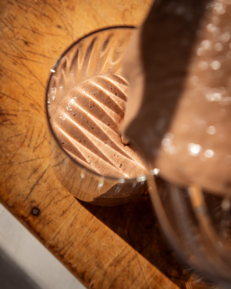 An overhead shot shows a chocolaty smoothie being poured into a fluted glass. The photo is taken in direct sun light and the fluted texture of the glass shows as stripes across the top of the smoothie's surface.