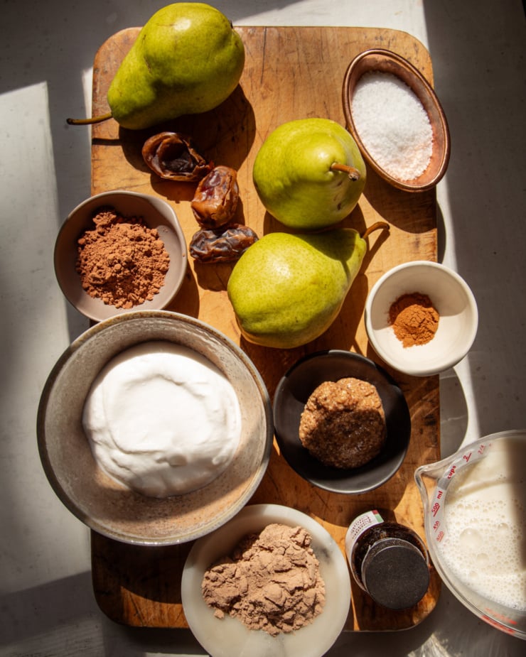 An overhead shot shows all ingredients needed to make a vegan chocolate pear smoothie. All ingredients are on a rough wooden board in direct sun light with heavy, dramatic shadows.