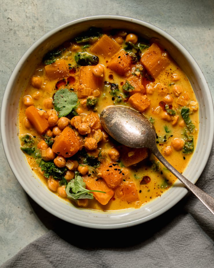 An overhead shot shows a bowl of butternut squash stew with coconut milk, kale and chickpeas. The broth is creamy and golden with bits of chopped cilantro garnishing the top. A spoon is sticking out of the bowl. This portion is also garnished with some chili crisp.