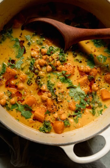 An overhead shot shows a pot of butternut squash stew. A wooden ladle is sticking out of the pot. The broth appears creamy and golden.