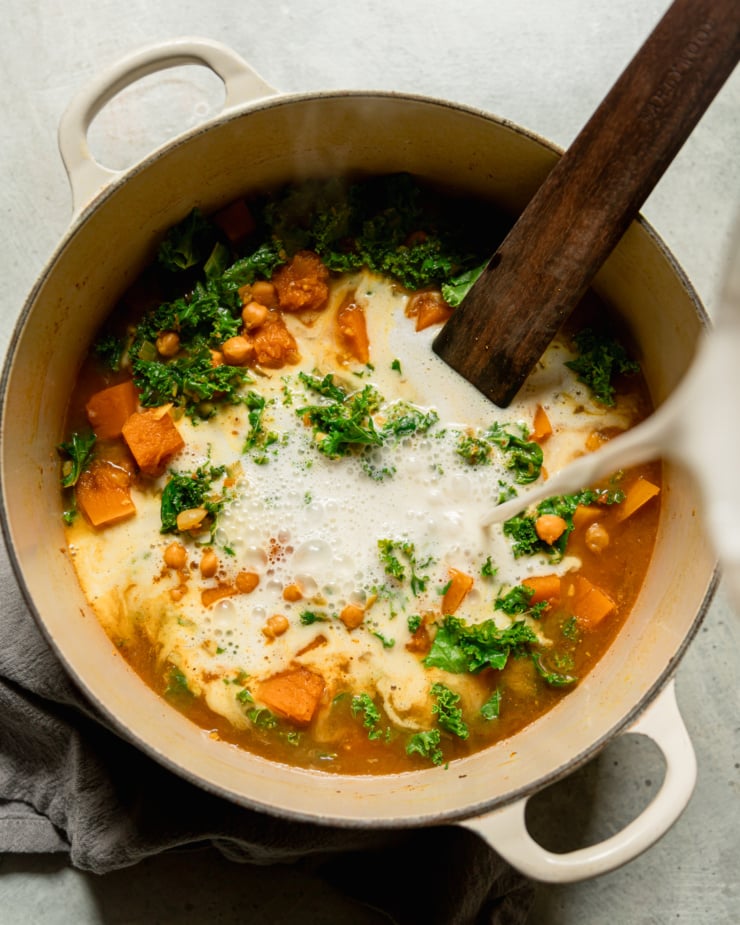 An overhead shot shows coconut milk being poured into a pot with wilted kale, cubed squash, chickpeas, and broth. A wooden stirring utensil is sticking out of the pot.