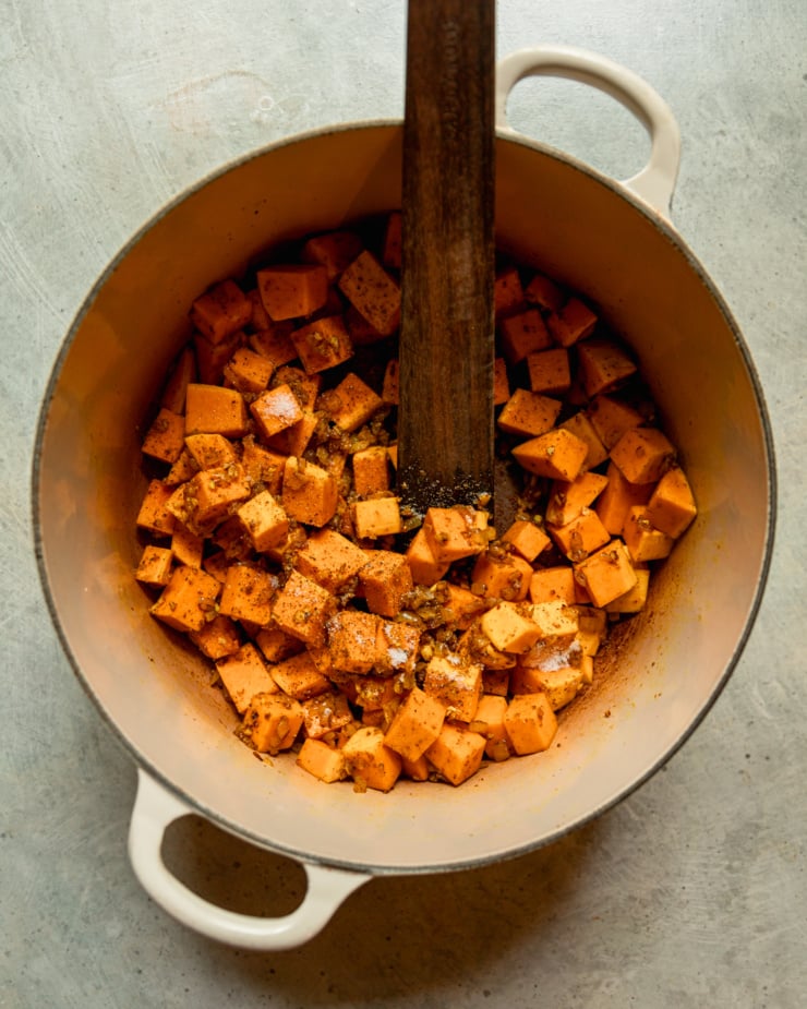 An overhead shot shows cubes of squash being sautรฉed in a big pot with spices. A wooden stirring utensil is sticking out of the pot.