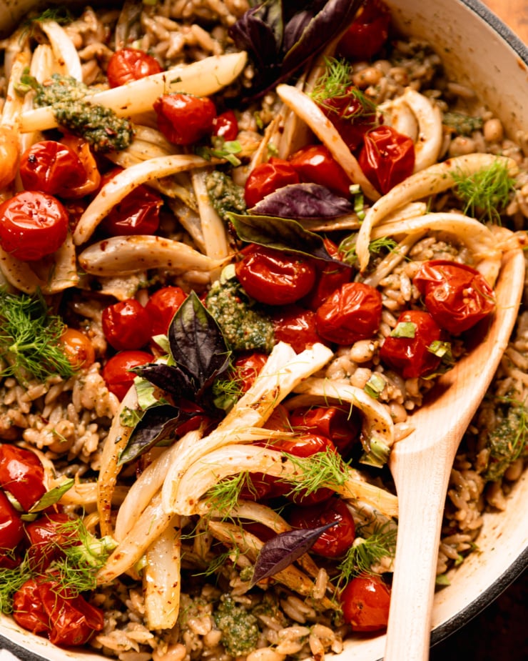 An up close, overhead shot shows a pesto orzo dish topped with roasted cherry tomatoes, fennel, fresh basil, and fennel fronds. A wooden serving spoon is sticking out of the braiser pot that everything is contained in.