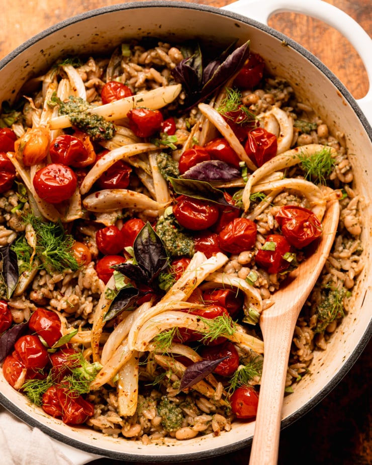 An up close, overhead shot shows a pesto orzo dish topped with roasted cherry tomatoes, fennel, fresh basil, and fennel fronds. A wooden serving spoon is sticking out of the braiser pot that everything is contained in.
