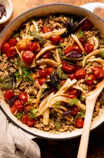 An overhead shot shows a pesto orzo dish topped with roasted cherry tomatoes, fennel, fresh basil, and fennel fronds. A wooden serving spoon is sticking out of the braiser pot that everything is contained in.