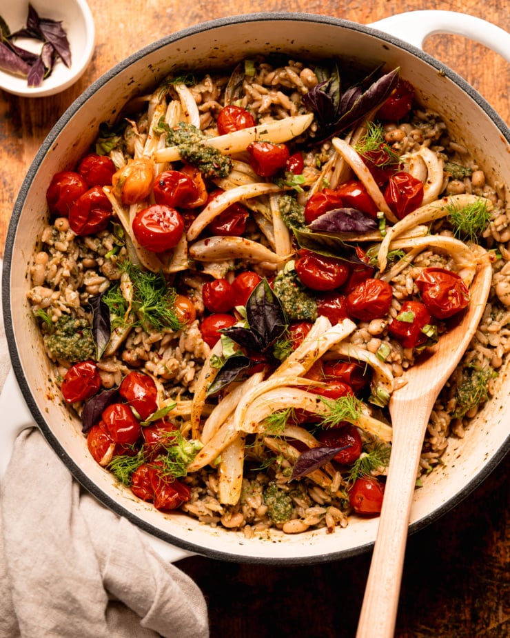An overhead shot shows a pesto orzo dish topped with roasted cherry tomatoes, fennel, fresh basil, and fennel fronds. A wooden serving spoon is sticking out of the braiser pot that everything is contained in.