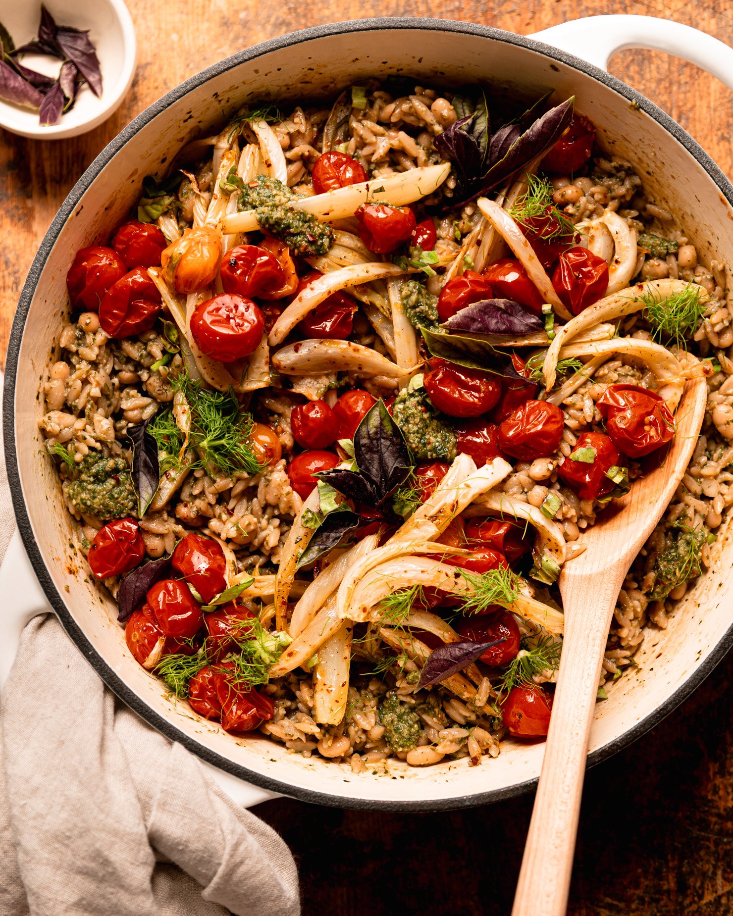 An overhead shot shows a pesto orzo dish topped with roasted cherry tomatoes, fennel, fresh basil, and fennel fronds. A wooden serving spoon is sticking out of the braiser pot that everything is contained in.